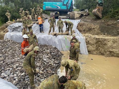 About 20 soldiers and some civilians working in flooded area with plastic, rocks and a digger